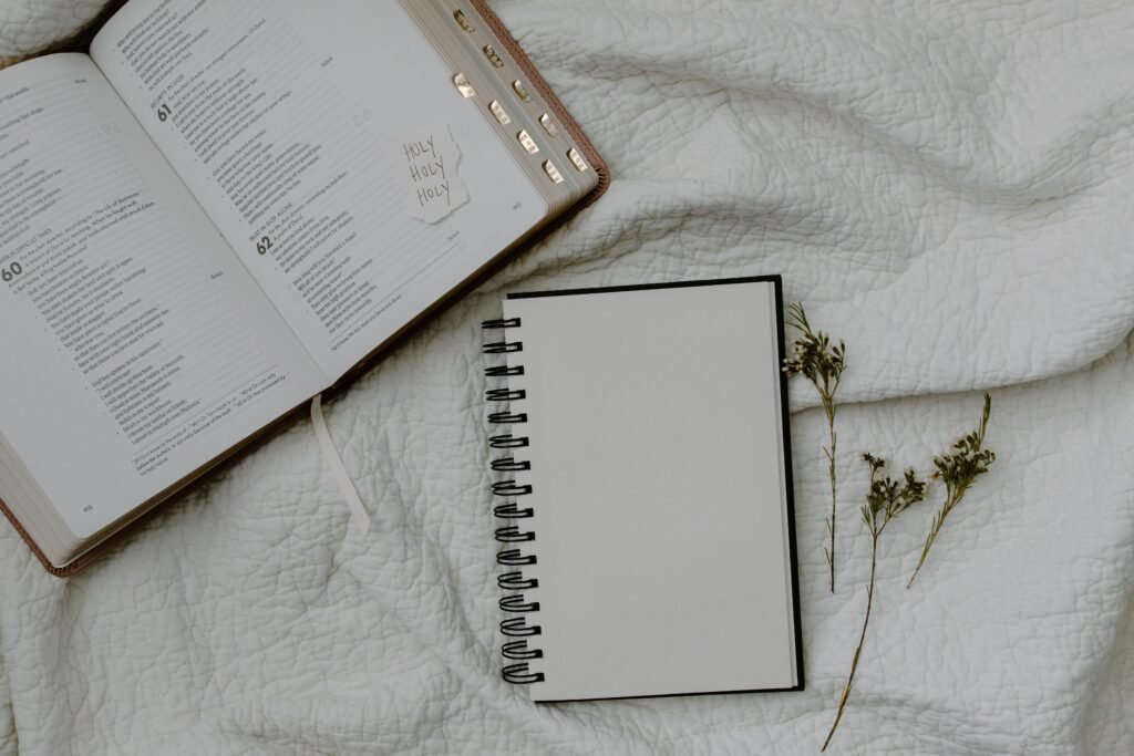 Top view of an open Bible, a blank notebook, and dried flowers on a textured white cloth, perfect for spiritual or study themes.