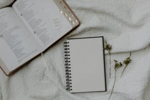 Top view of an open Bible, a blank notebook, and dried flowers on a textured white cloth, perfect for spiritual or study themes.